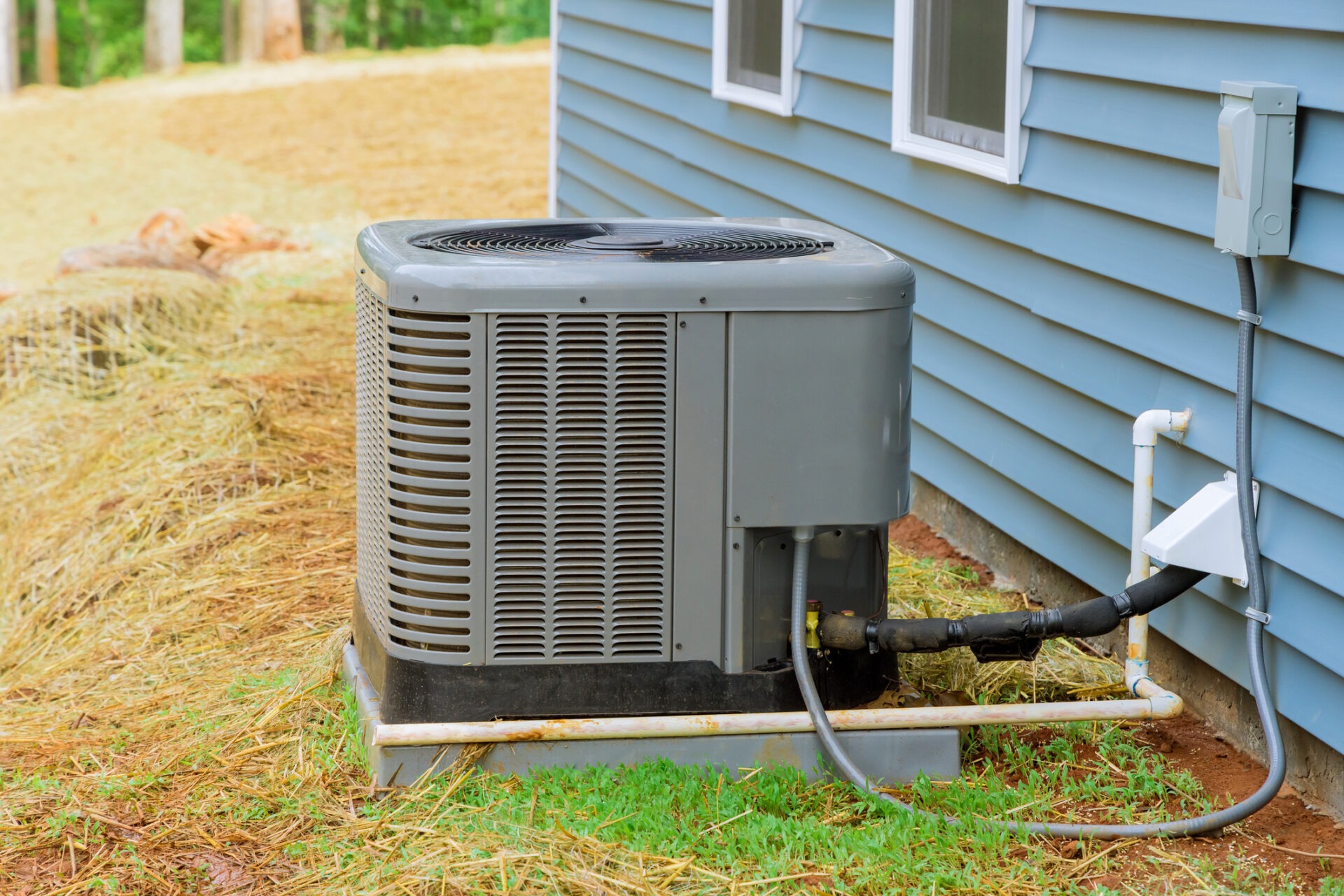 An outdoor central air conditioning unit positioned next to a blue-sided house with visible electrical and refrigerant lines on a grassy base.