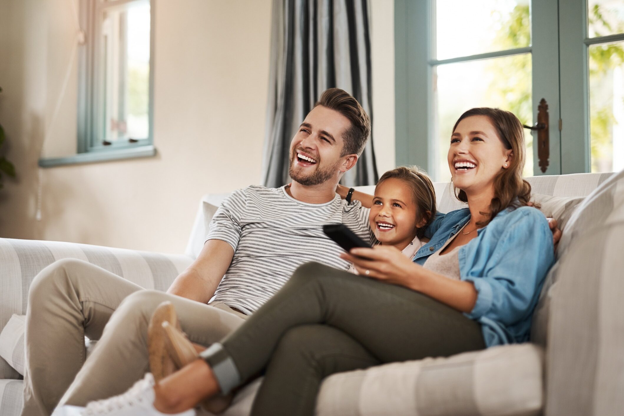 Two adults and a child are sitting on a sofa, smiling and laughing together. One person is holding a remote, suggesting they are watching TV.
