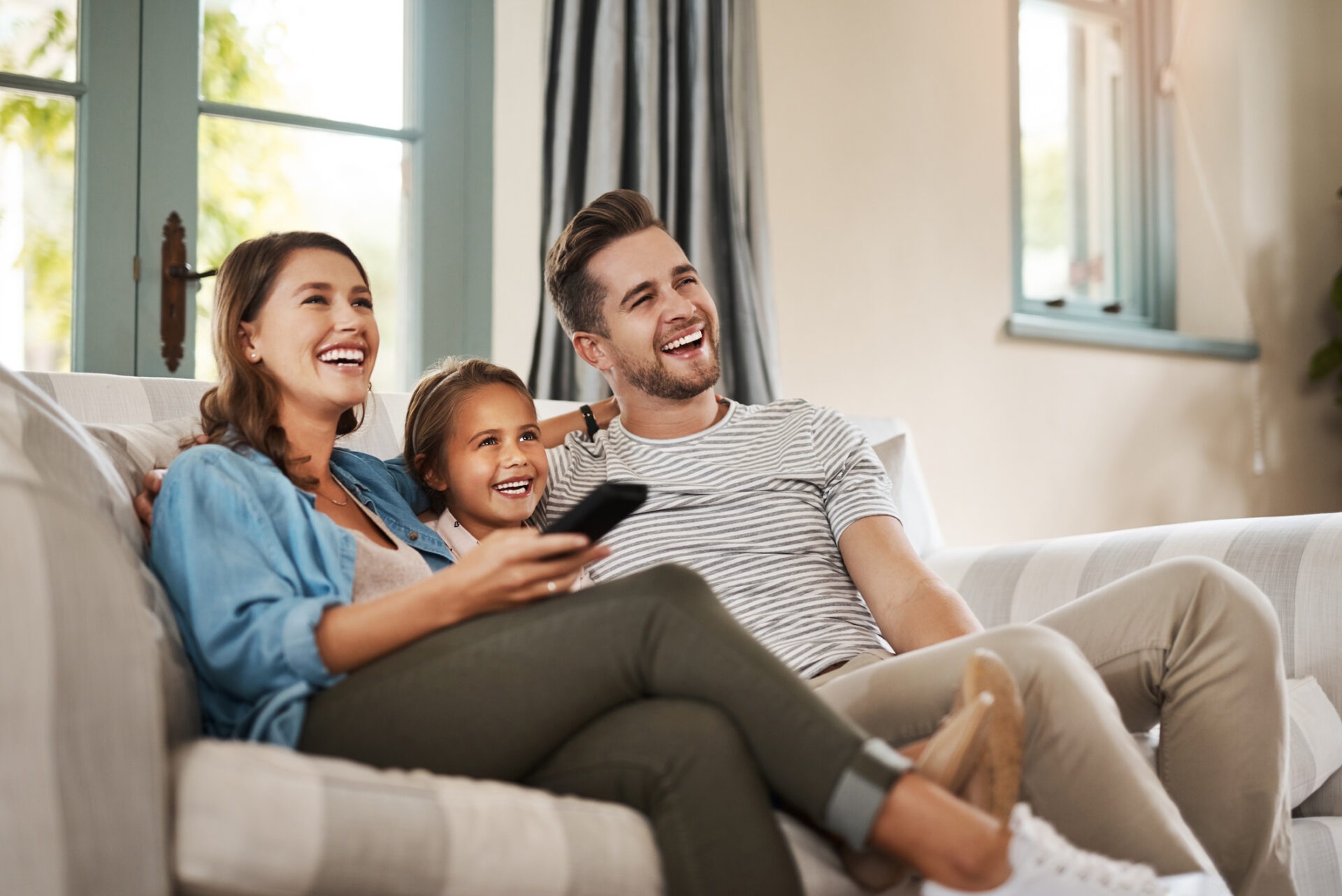 Two adults and a child are sitting on a couch, smiling and looking to the side, while one person holds a remote control. They appear happy.
