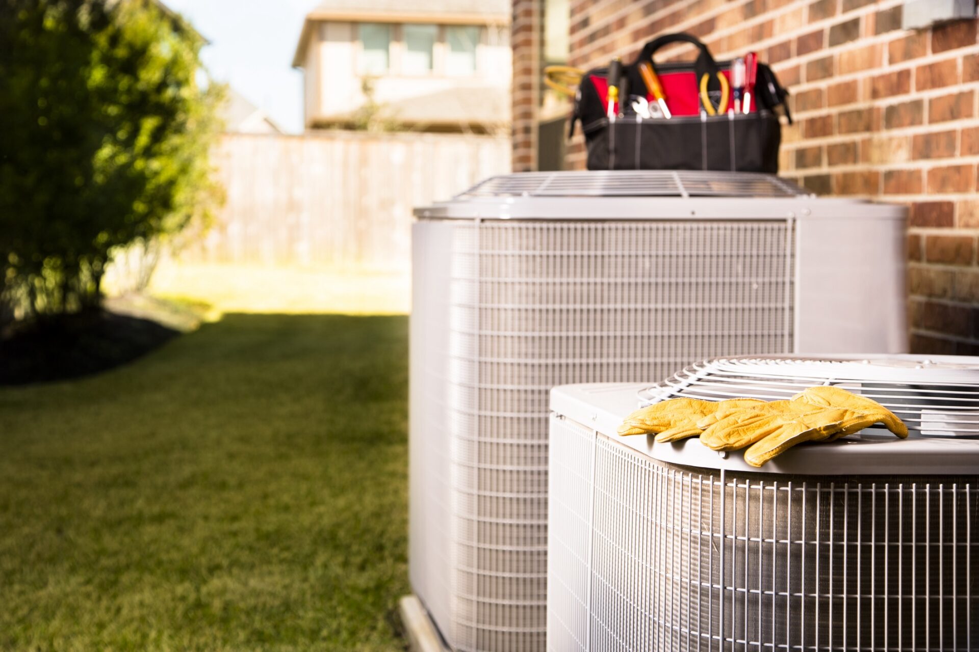 Two air conditioning units outside a house with a pair of yellow gloves on one and a tool bag in the background, suggesting maintenance work.