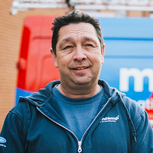 A person is standing outdoors wearing a blue jacket with a logo, slightly smiling at the camera. A red brick building and a blue truck are in the background.