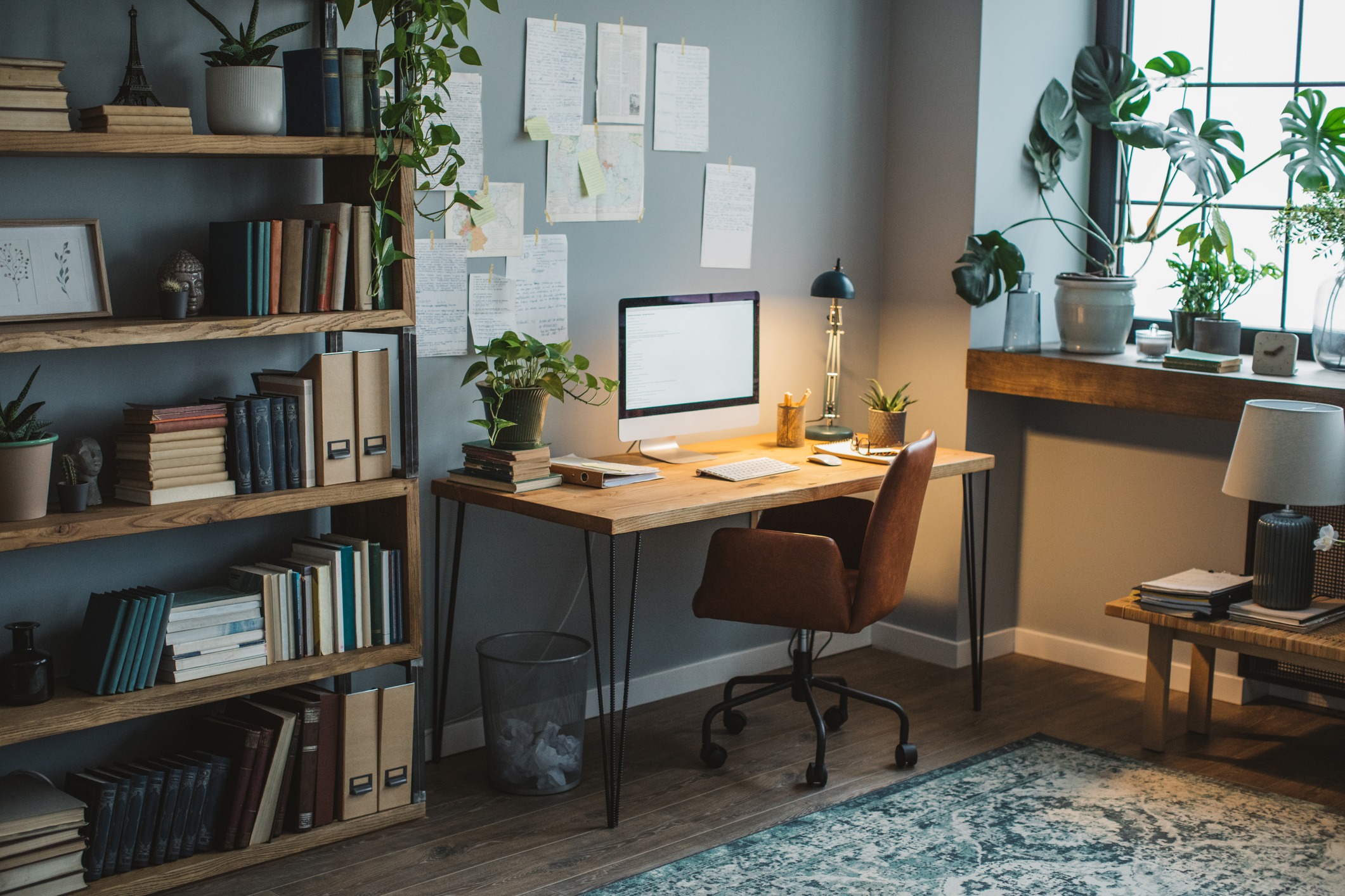 A cozy home office with wooden shelves, books, a desk, computer, decorations, plants, and papers on the wall; warm lighting and a patterned rug.