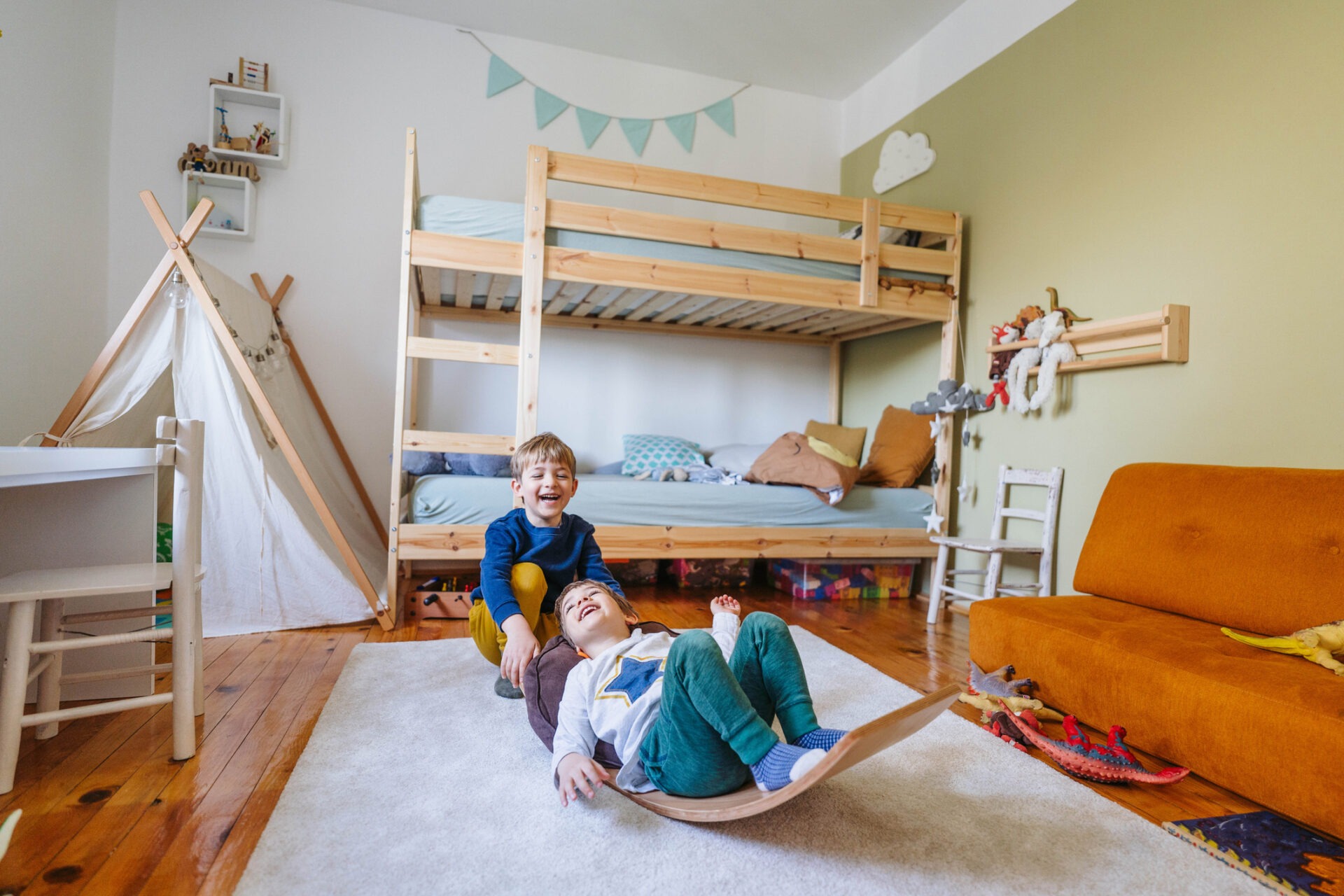 Two smiling children play in a colorful room with a bunk bed, teepee, toys, and a mustard-colored couch. A cozy and playful atmosphere.