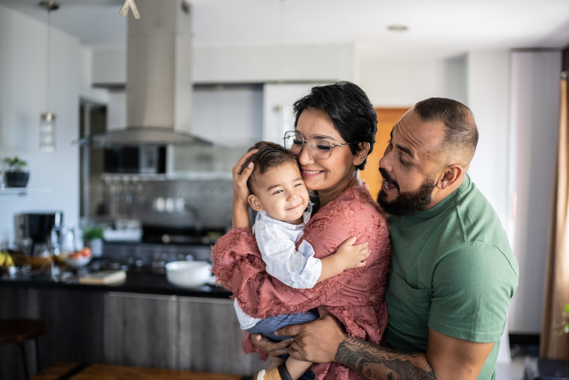 A family of three—a child and two adults—are embracing warmly in a well-equipped home kitchen, exuding happiness and affection.