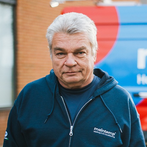 A person with grey hair and a serious expression is standing outdoors. They are wearing a navy blue jacket with the "maintemp" logo on it.