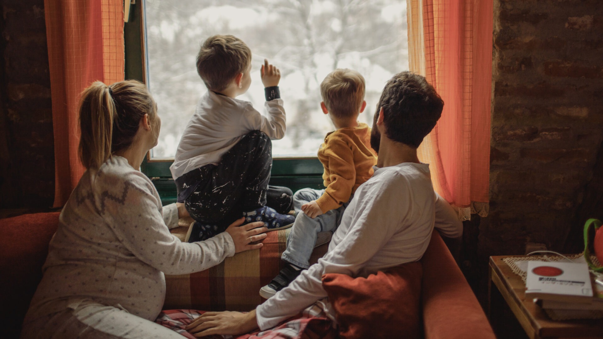 Two adults and two children sit near a window, gazing outside at a snow-covered landscape, with warm lighting and cozy interior surroundings.