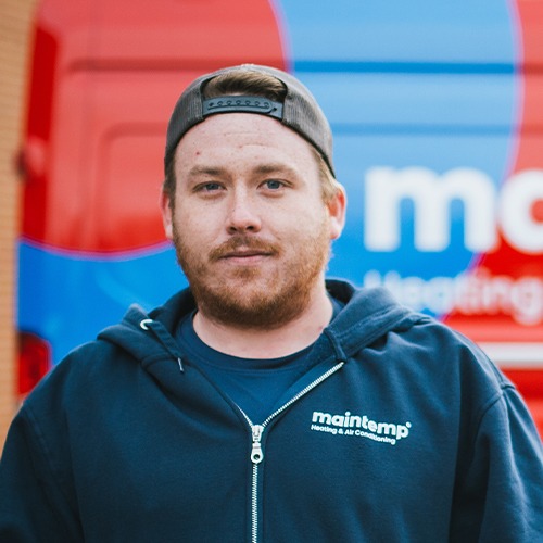 A person wearing a dark hoodie and cap stands in front of a red vehicle with "maintemp" printed on it, looking directly at the camera.