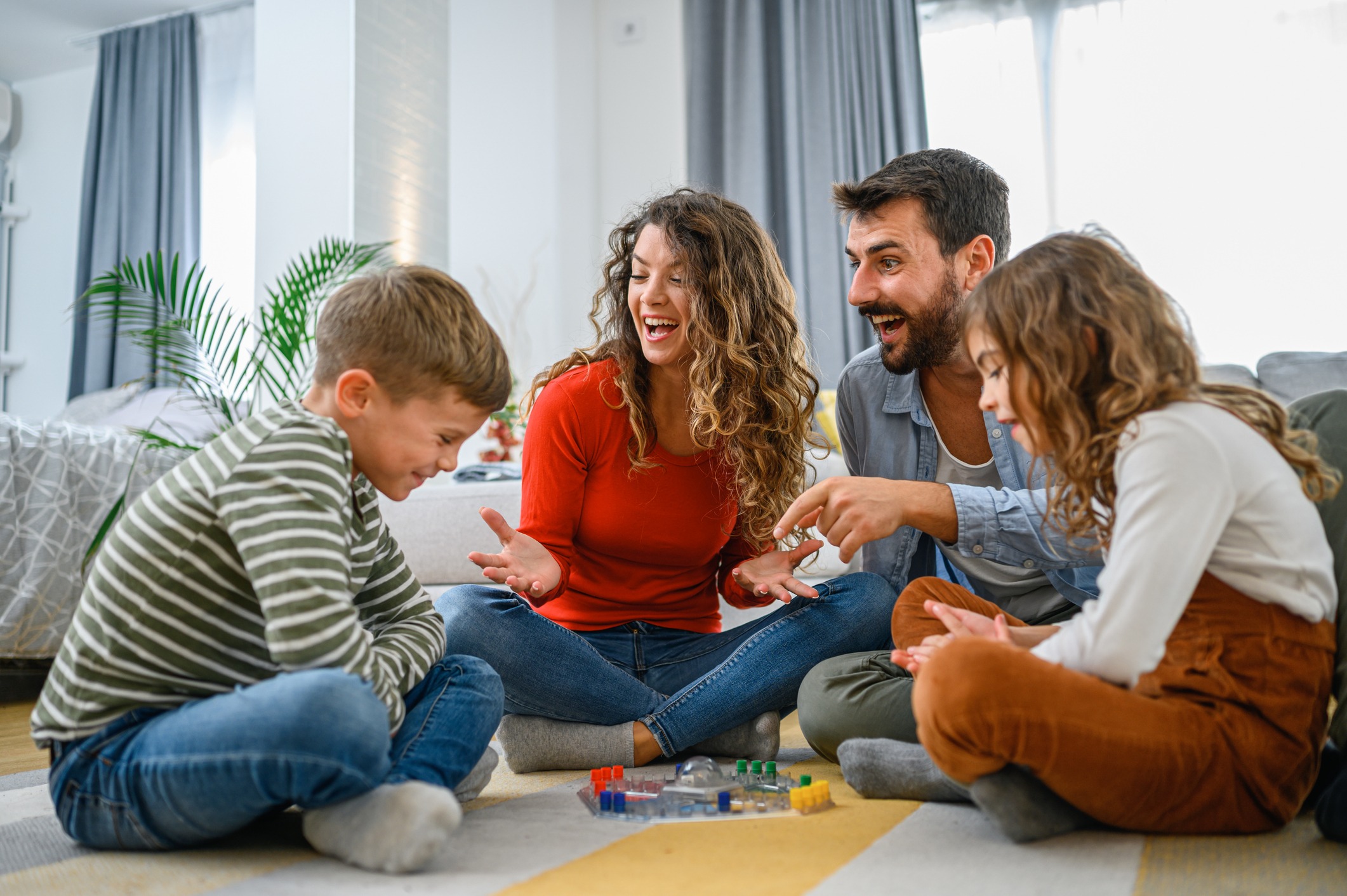A family is laughing and playing a board game together on the floor in a bright, cozy living room setting.