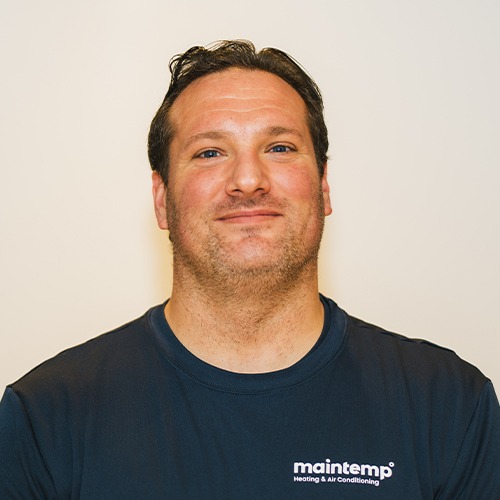 A person is smiling against a white background, wearing a dark blue shirt with "maintemp Heating & Air Conditioning" logo, short dark hair, and stubble.