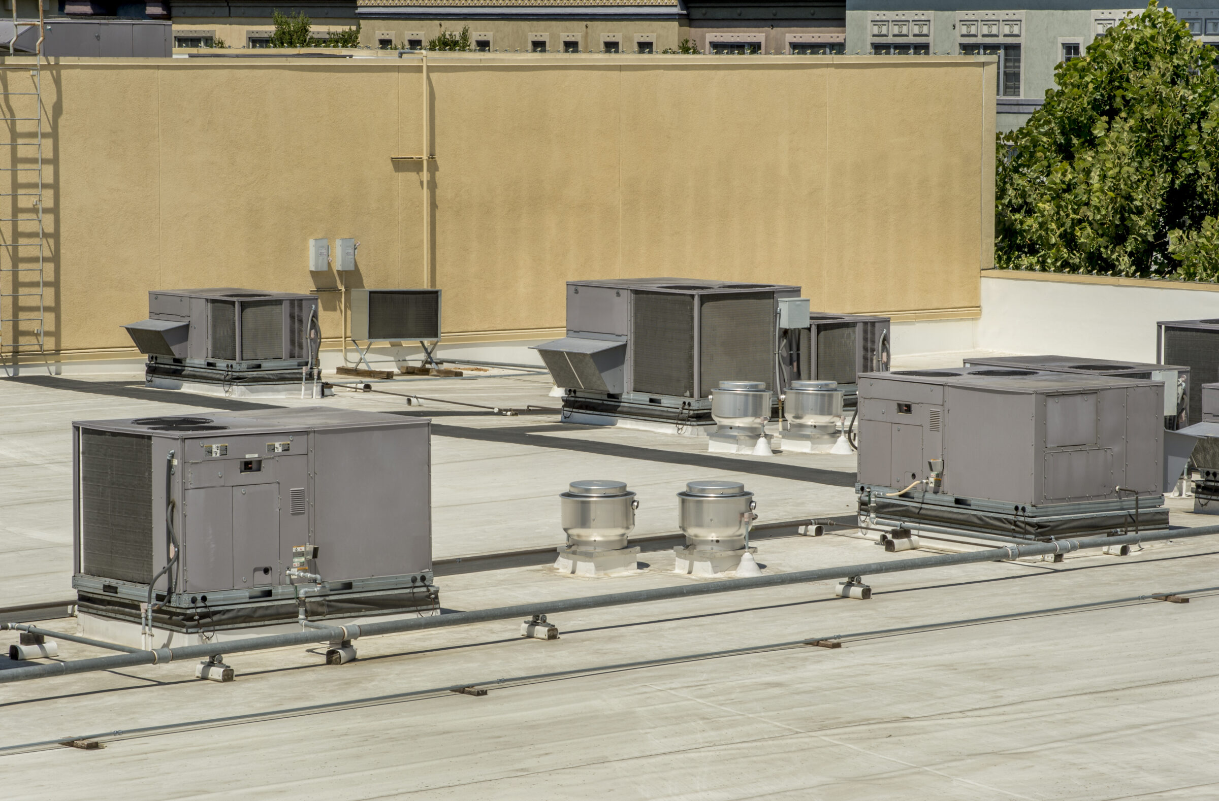 This image shows a flat rooftop with multiple HVAC units, vent pipes, and a beige wall in the background. No persons are visible.