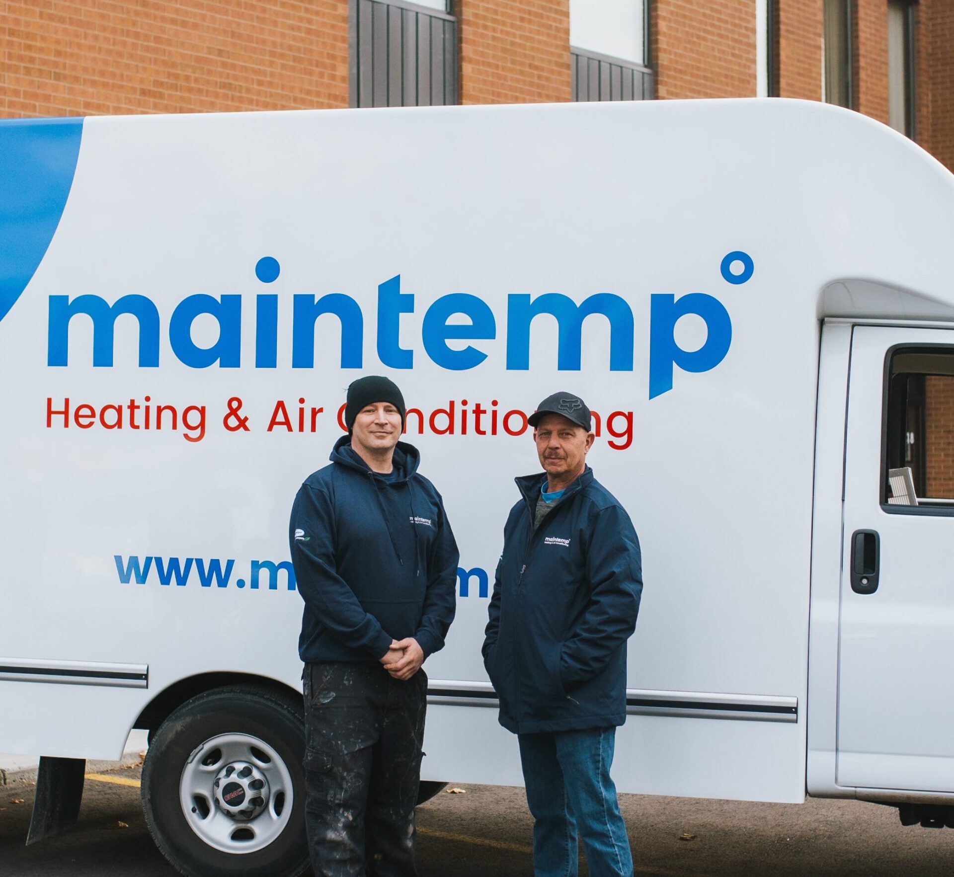 Two people are standing in front of a white van with "maintemp Heating & Air Conditioning" branding, wearing company jackets, smiling, and looking at the camera.