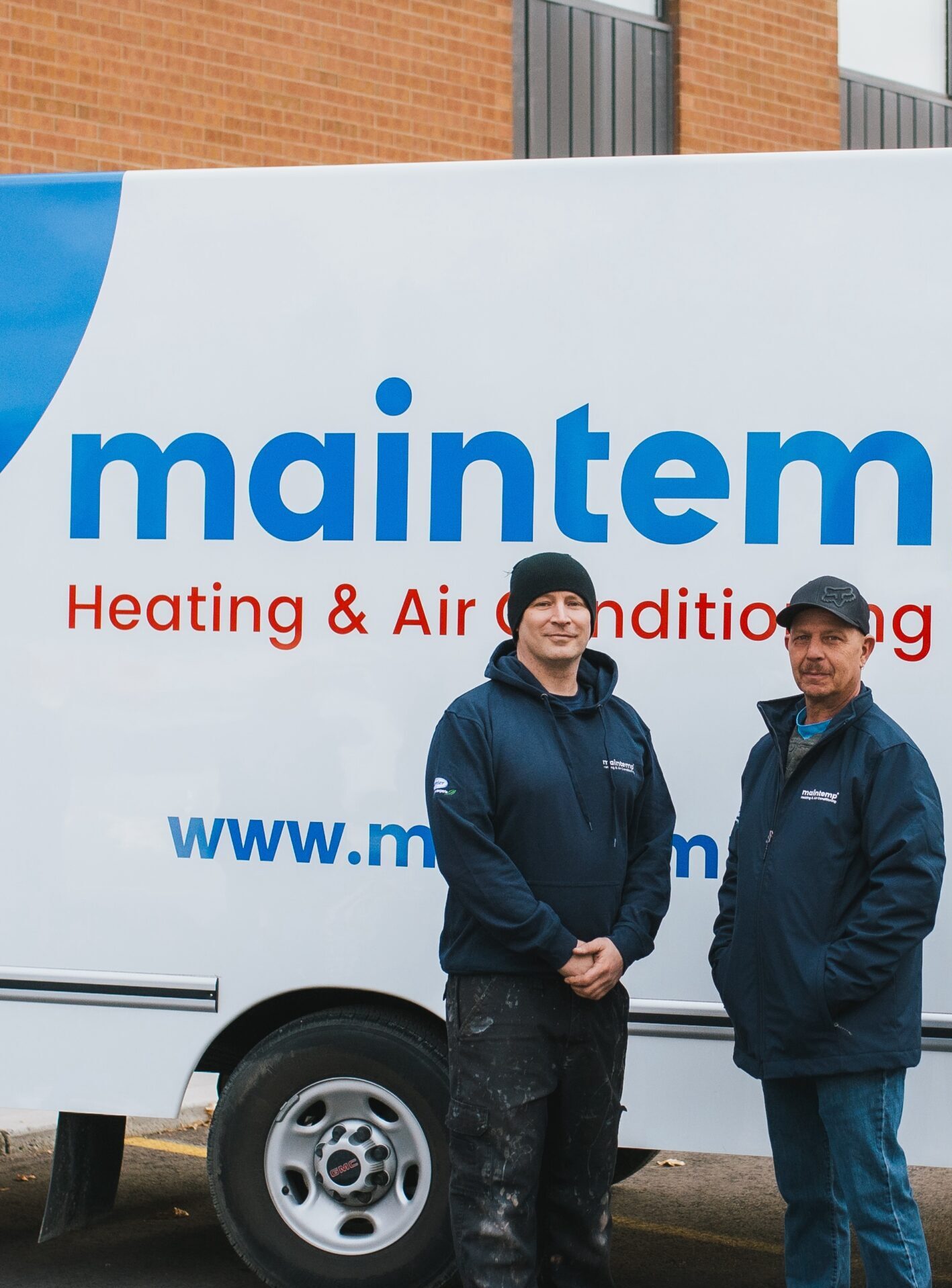 Two people stand in front of a white service van with "maintemp Heating & Air Conditioning" written on it, smiling slightly, in work attire.