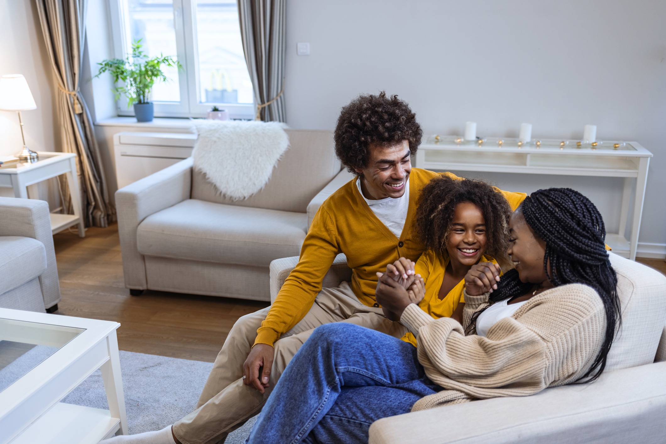 A happy family with two adults and a child is laughing together on the floor by a couch in a cozy living room setting.