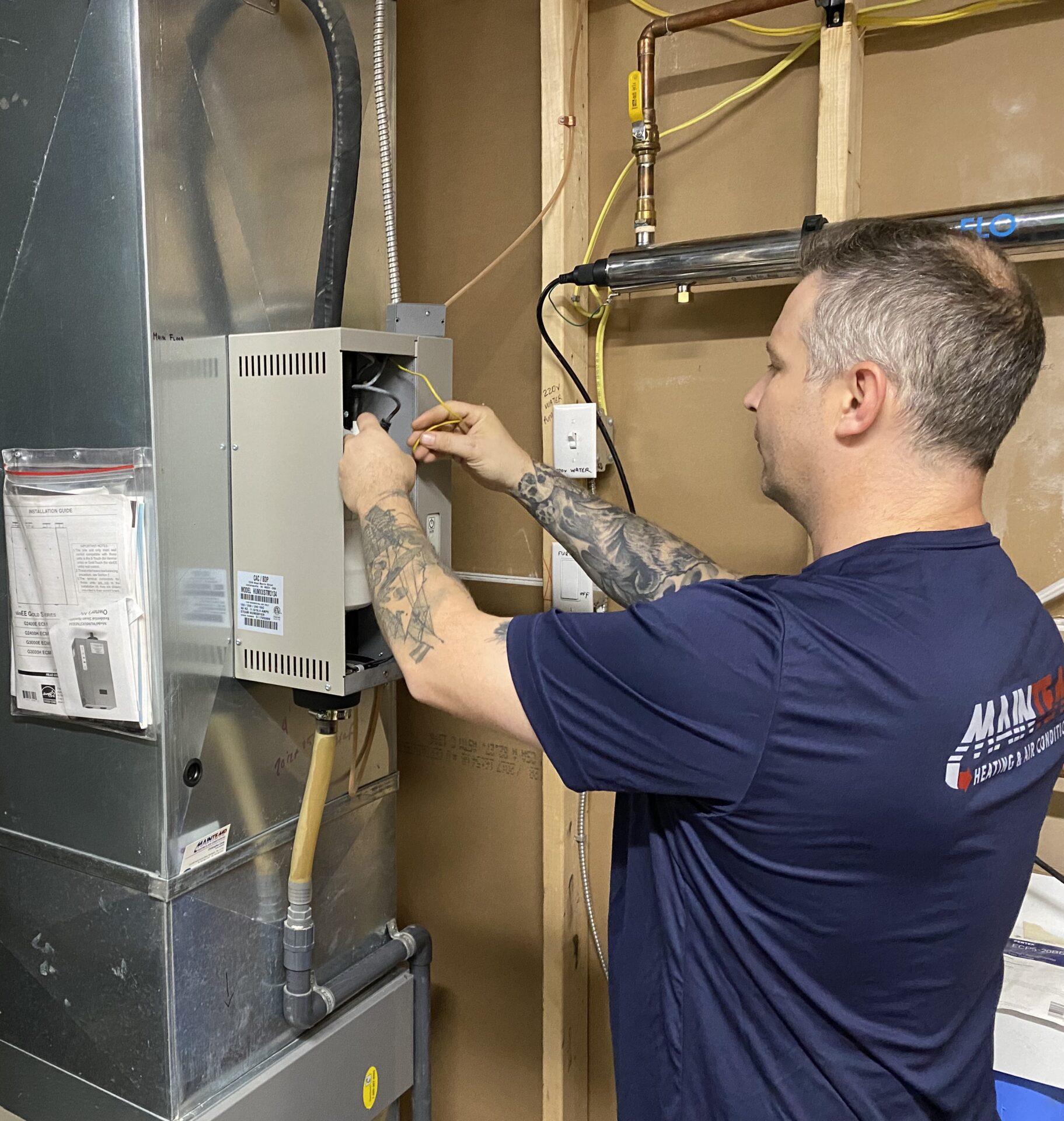 A person with tattoos is working on electrical components within a heating ventilation and air conditioning (HVAC) unit in a utility space.