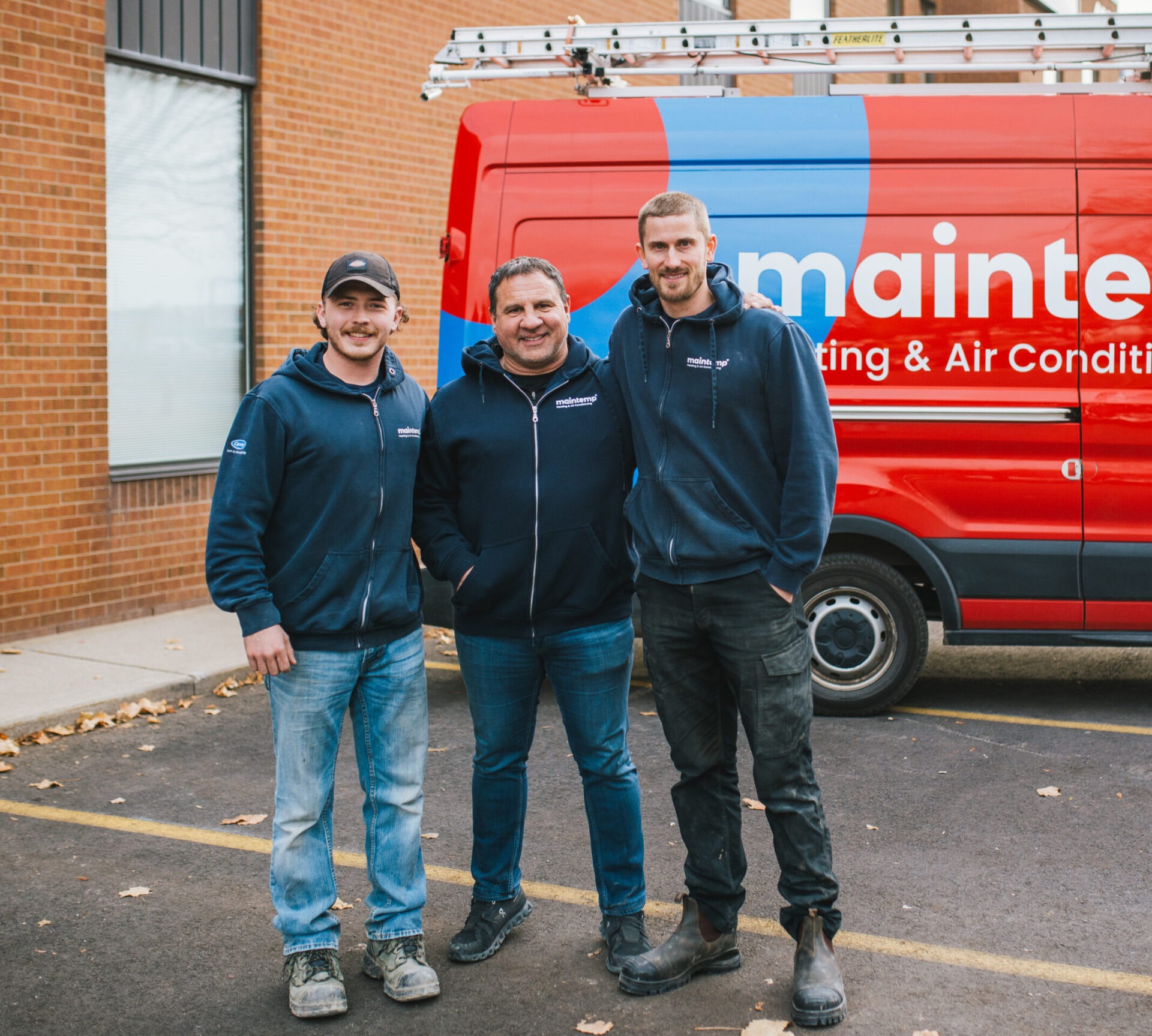 Three people in matching company jackets stand smiling in front of a red service van with "maintenance" text, suggesting they work in HVAC services.