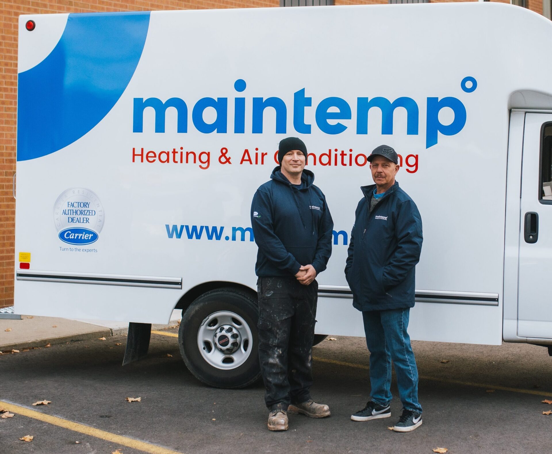 Two people are standing in front of a white van with "Maintemp Heating & Air Conditioning" logos, looking towards the camera with a brick building behind them.