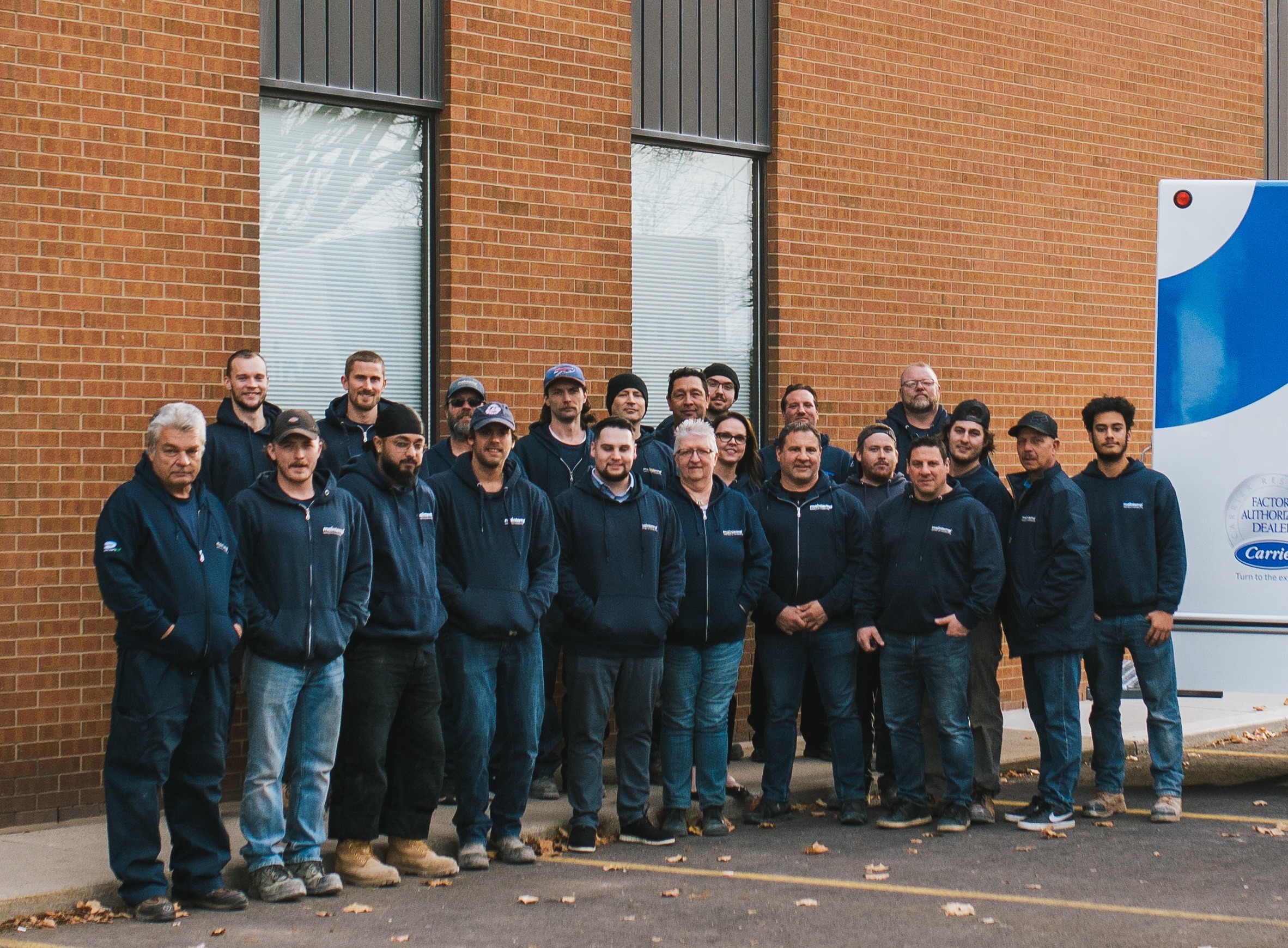 A group of people in matching uniforms posing together in front of a brown brick building, next to a vehicle with a company logo.