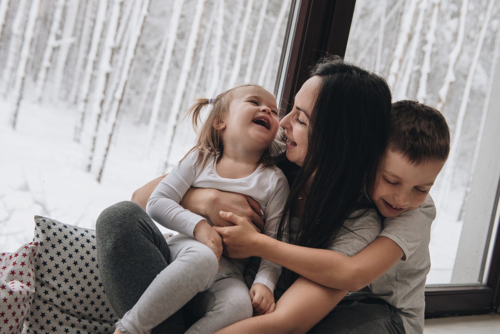 A person is sitting by a window, laughing and embracing two children. They appear happy and playful against a snow-covered forest backdrop.