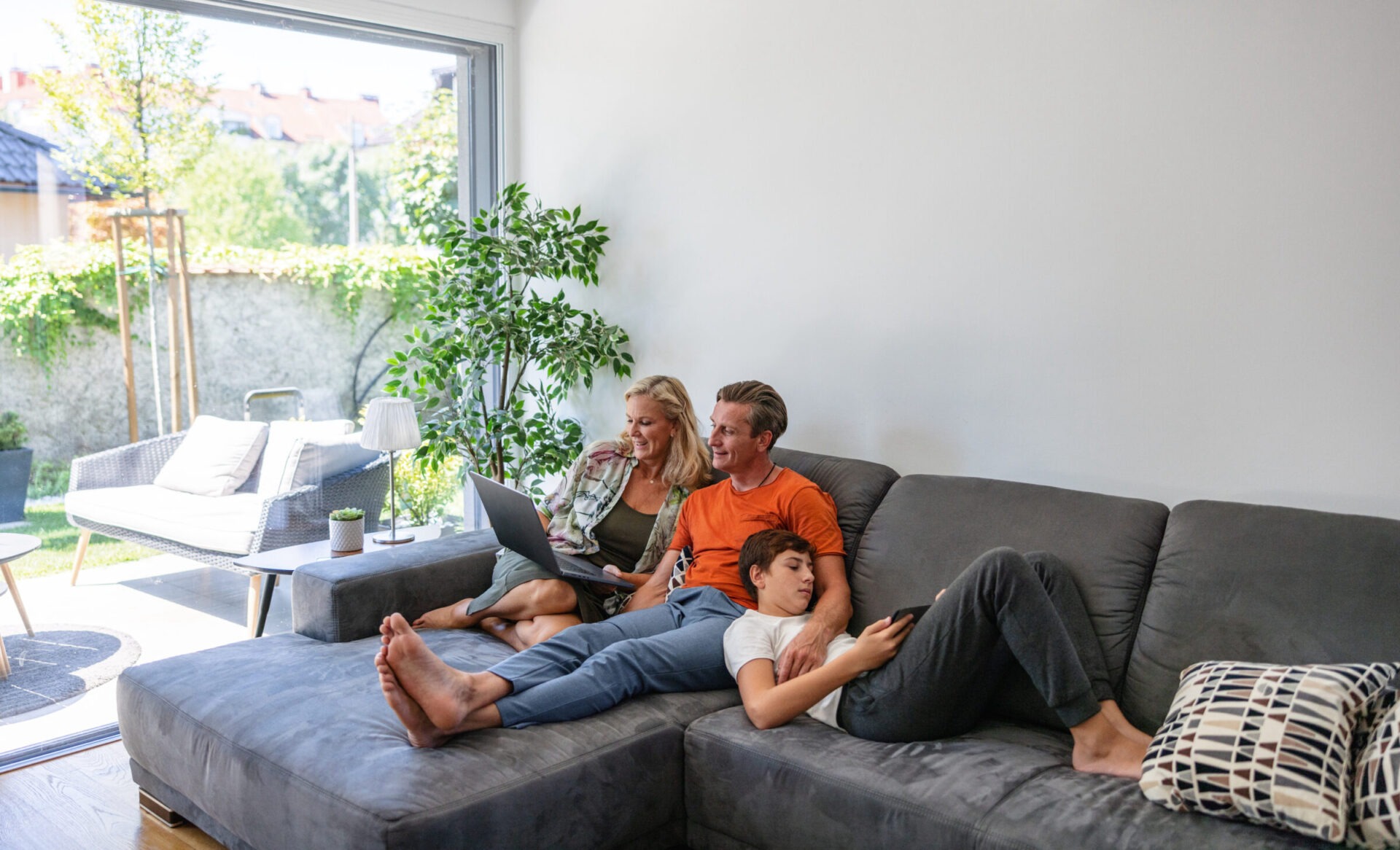 Two adults and a child are relaxing on a gray sofa in a bright living room, using a laptop and a smartphone, with garden view.