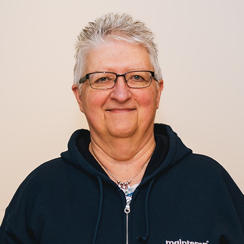 A person with short gray hair and glasses is smiling in front of a cream-colored background, wearing a navy blue hoodie with a visible nametag.