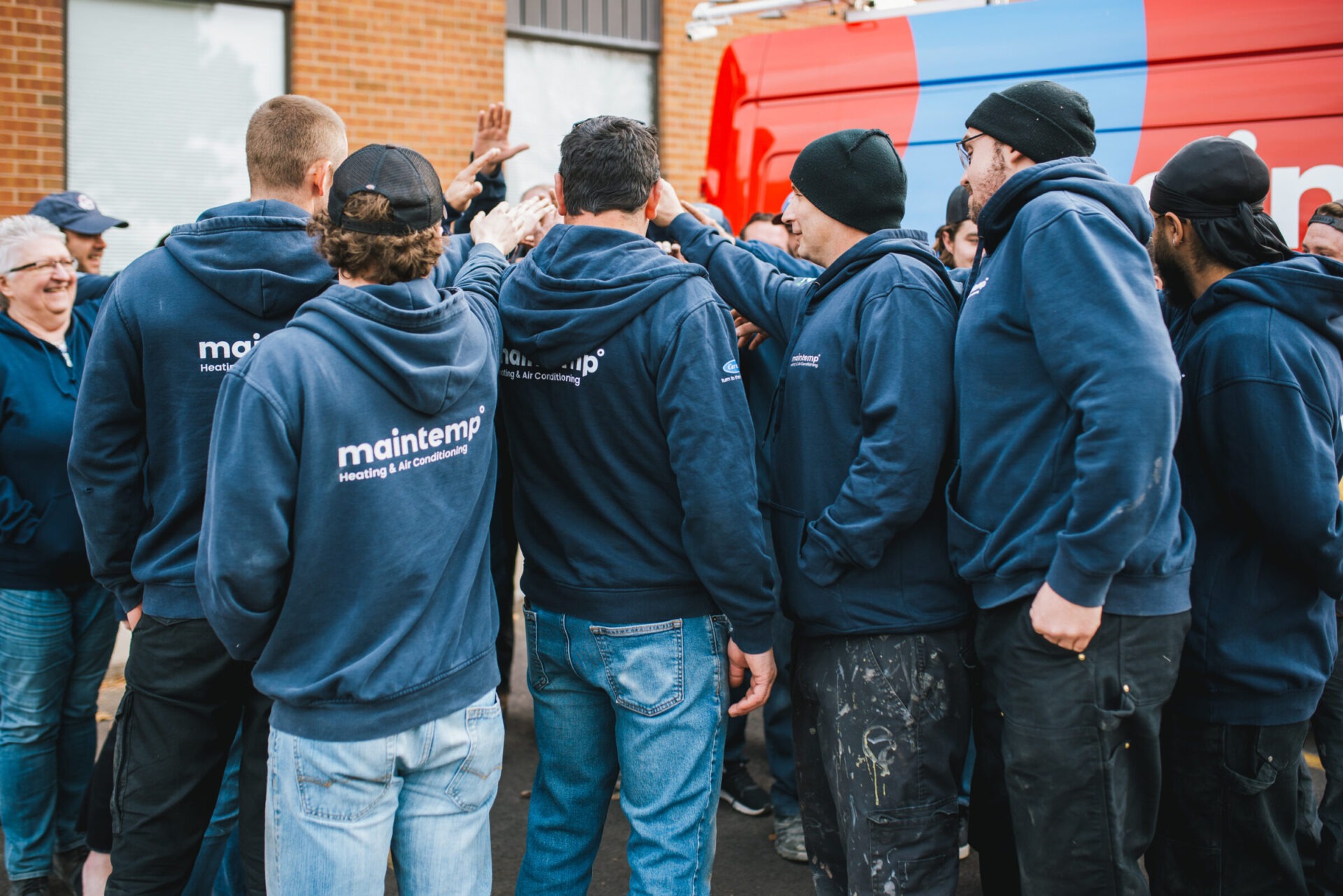 A group of people wearing matching navy blue hoodies with a company logo gathered outside, some with arms raised, possibly celebrating or team-building.