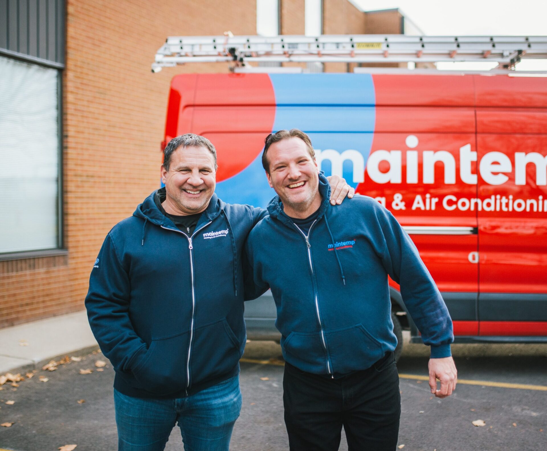 Two people smiling in front of a red van labeled "maintenance & Air Conditioning," wearing matching blue work jackets with logo, in daylight.