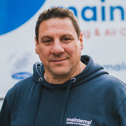 A person is wearing a blue hoodie with a "maintemp Heating & Air Conditioning" logo, smiling slightly, with a blurred company sign in the background.