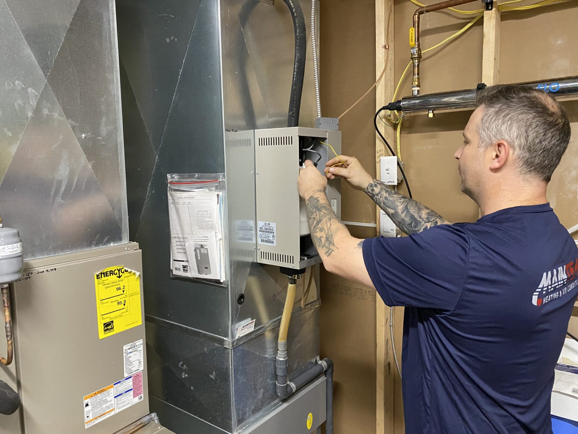 A person with tattoos is working on an HVAC system in a utility room, using a screwdriver. Visible equipment includes ductwork and water heaters.