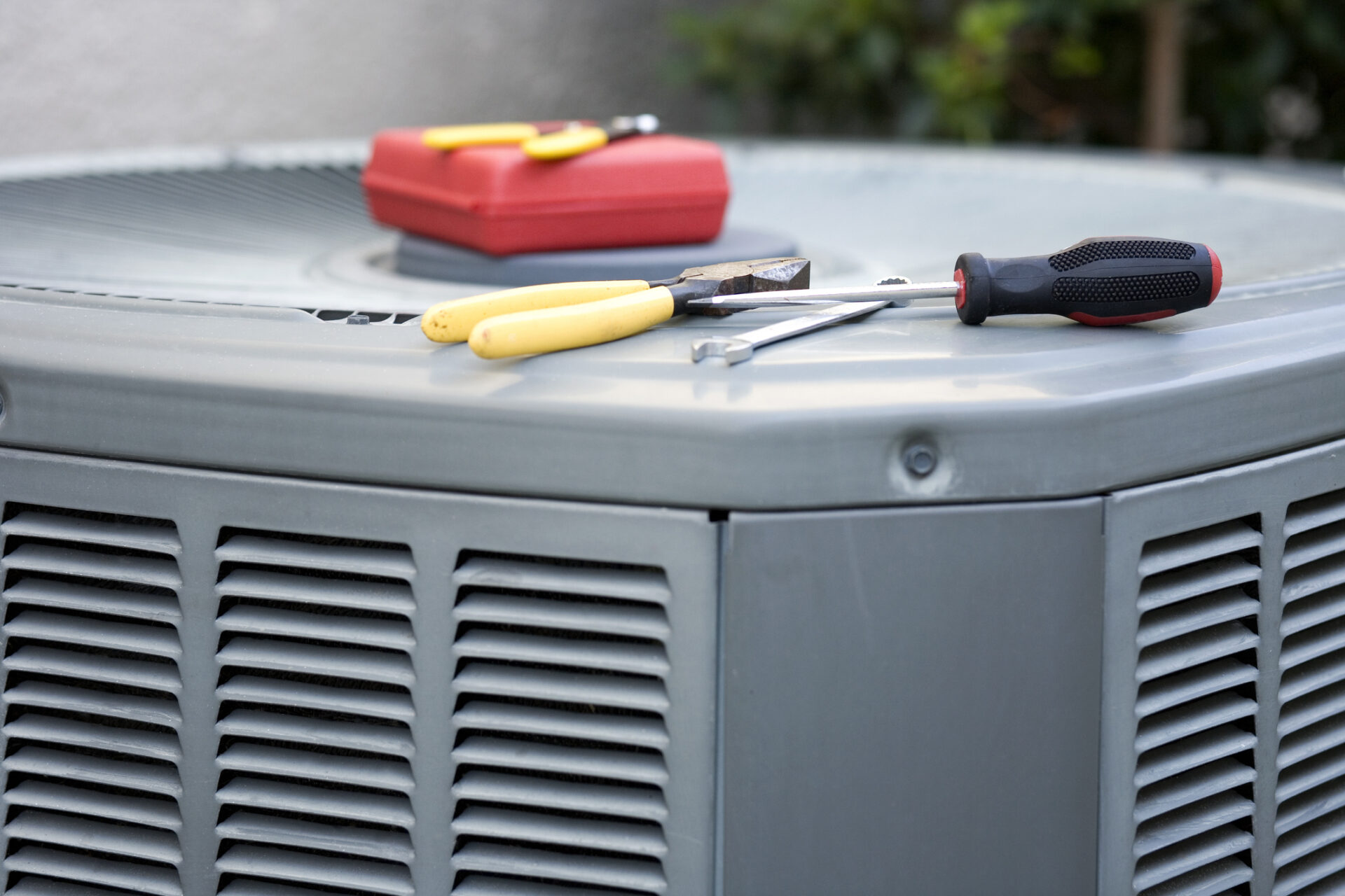 An air conditioning unit outdoors with tools on top, including pliers and a screwdriver, suggesting maintenance work, with a red toolbox nearby.