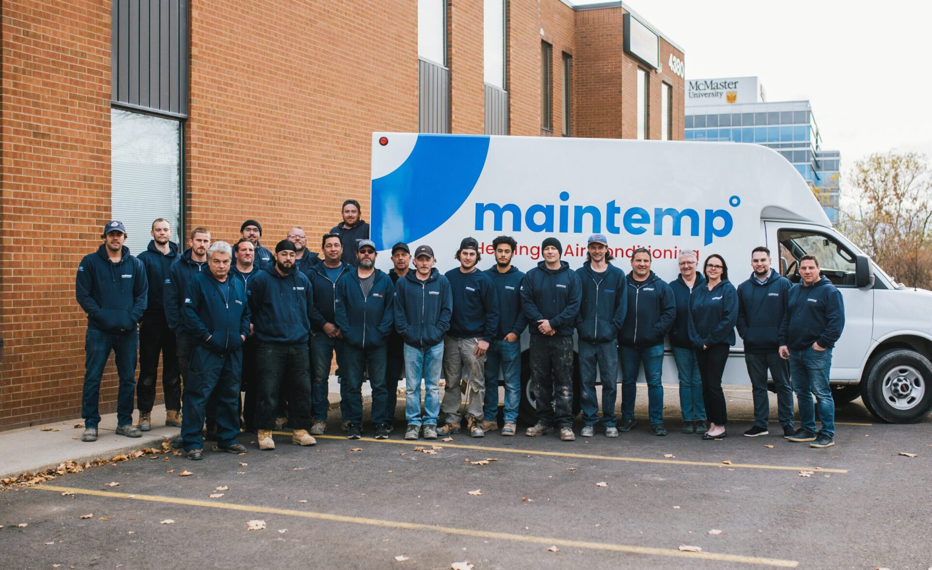 A group of twenty-two people in uniform stand in front of a commercial van with the "Maintemp" logo outside a building with the McMaster University sign.
