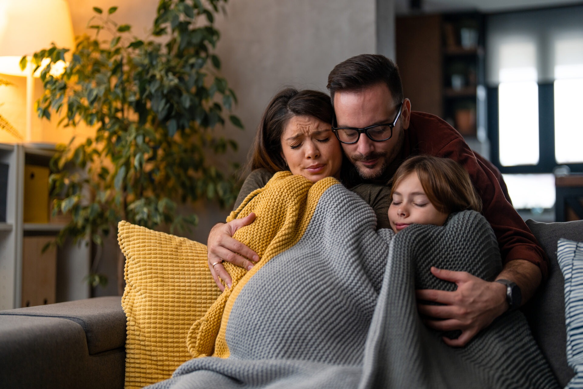 A person and two children are hugging on a couch, wrapped in blankets, with emotional, possibly sad expressions in a cozy room.