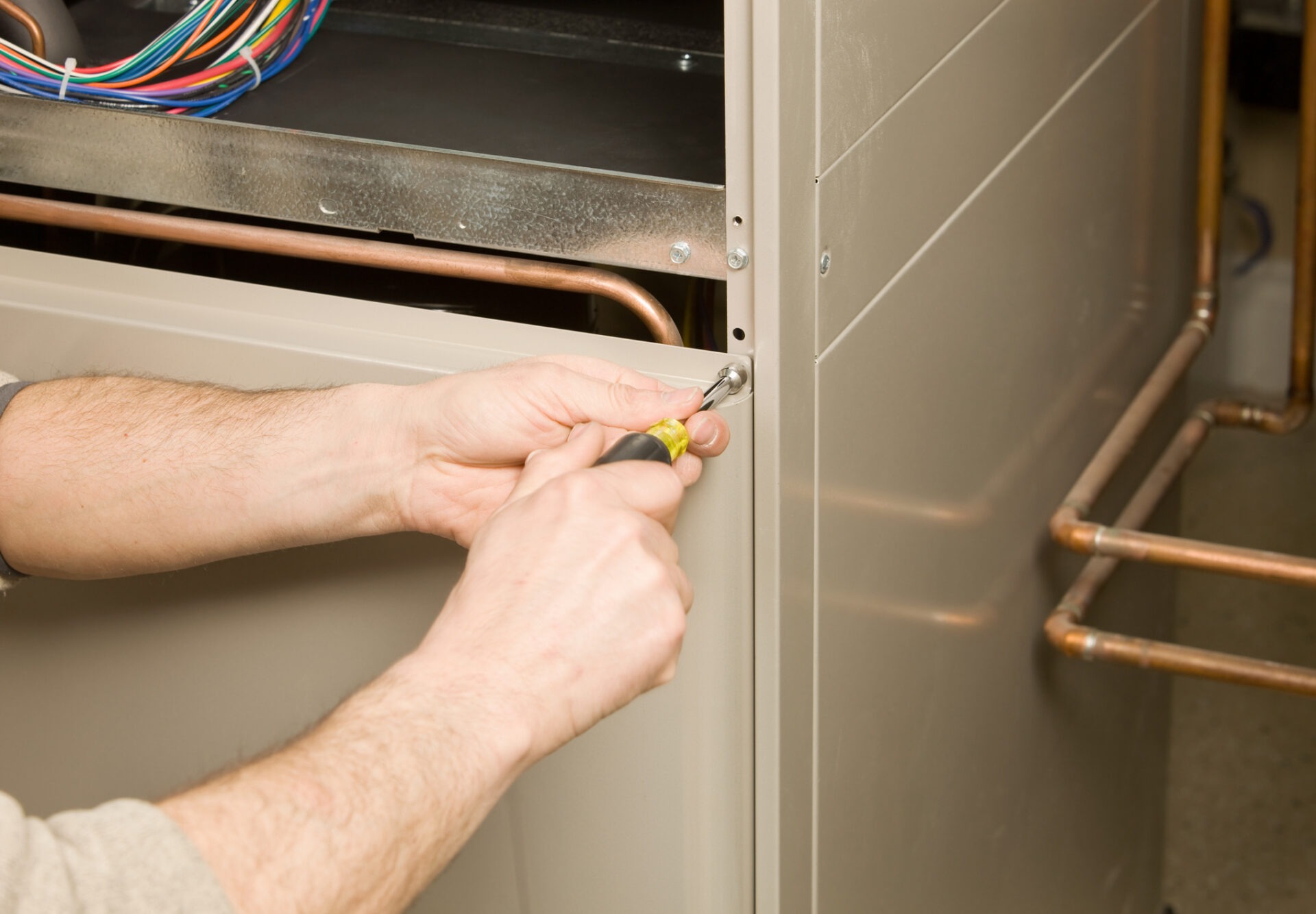 A person is using a screwdriver to work on a beige HVAC unit, with visible wires and copper pipes in the background.