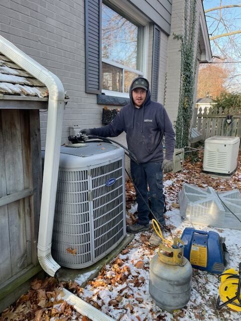 A person stands next to an outdoor air conditioning unit, surrounded by tools and a gas cylinder, indicating maintenance work, in a residential setting with snow.