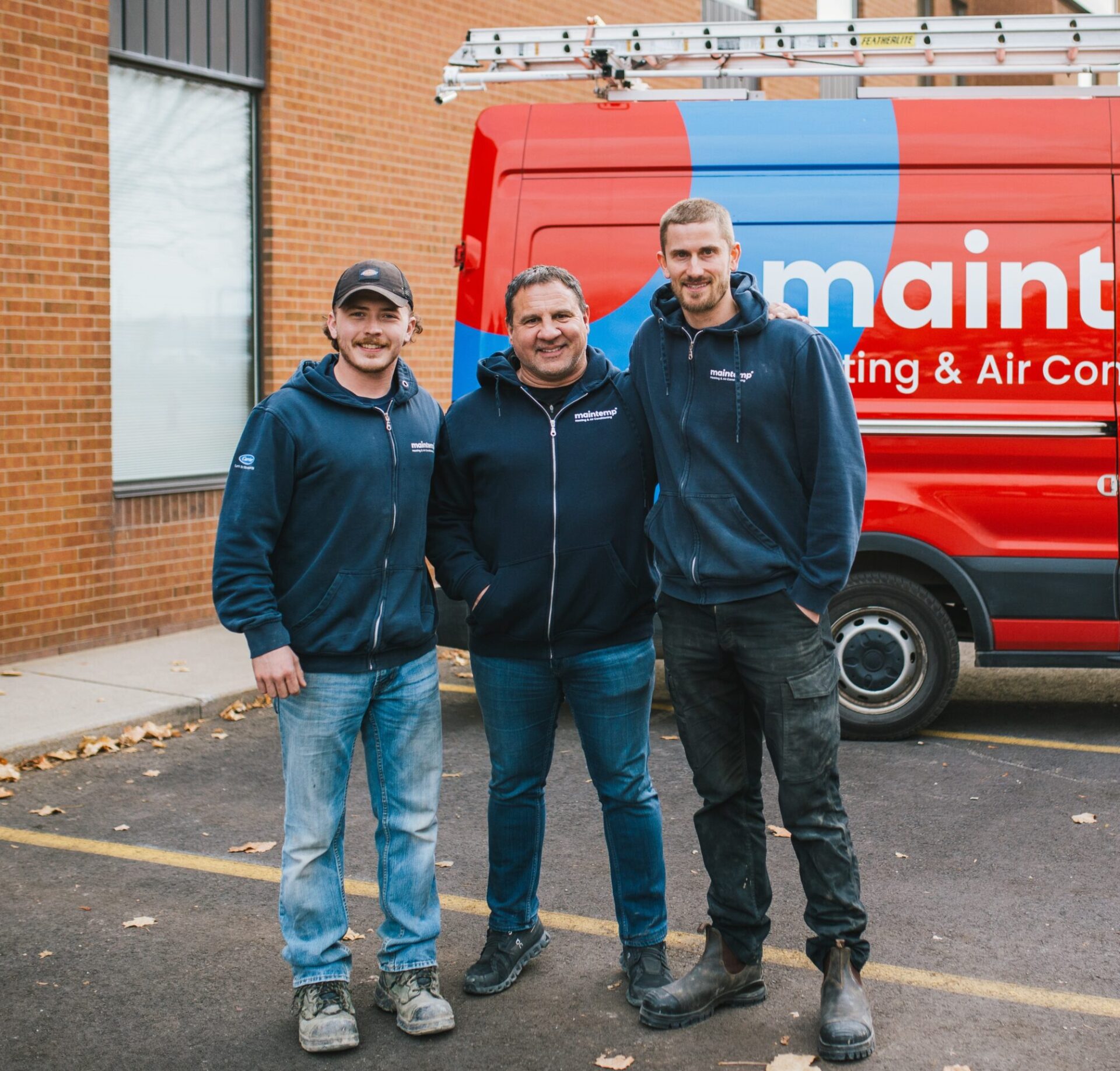 Three people wearing matching company jackets stand in front of a red service van with "maintenance" written on it, smiling for the camera.
