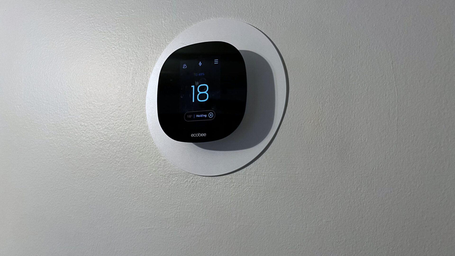 A modern, wall-mounted ecobee thermostat displays temperature at 18 degrees. The digital screen shows humidity and a holding temperature status.