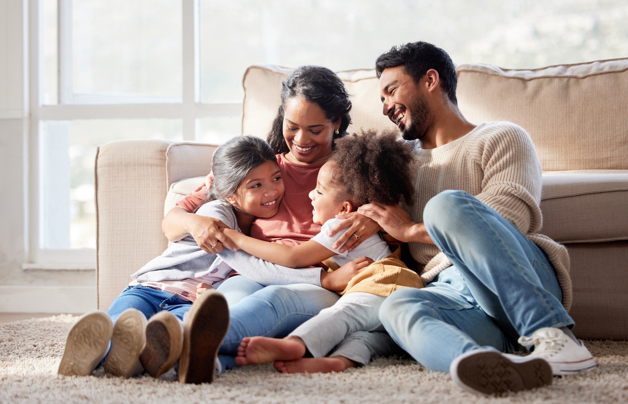 A happy family with two adults and two children is cuddled together on the floor, laughing in a sunlit room with a cozy ambiance.