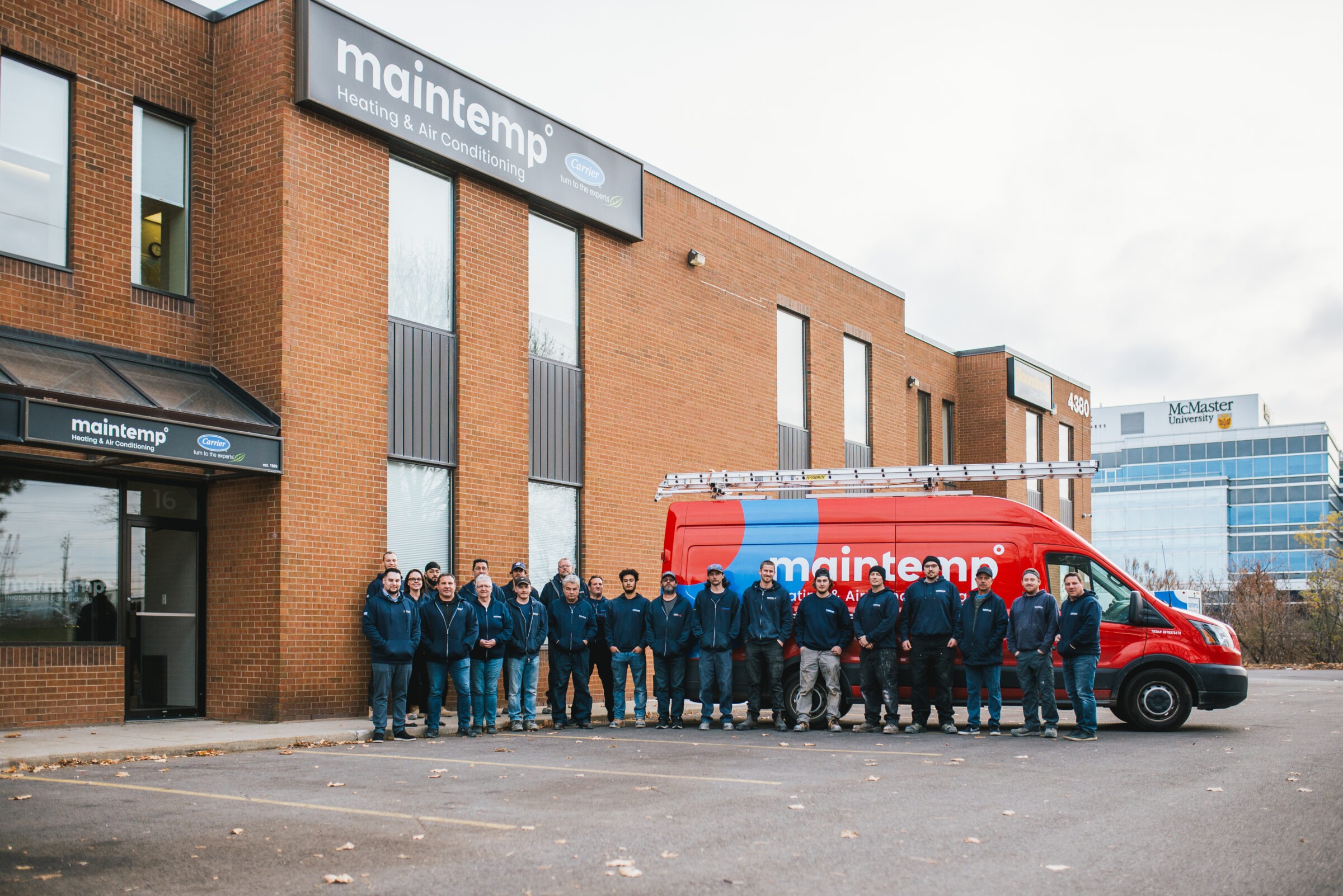 A group of people in matching blue jackets stands outside an industrial building beside a red van with "maintemp" logos.