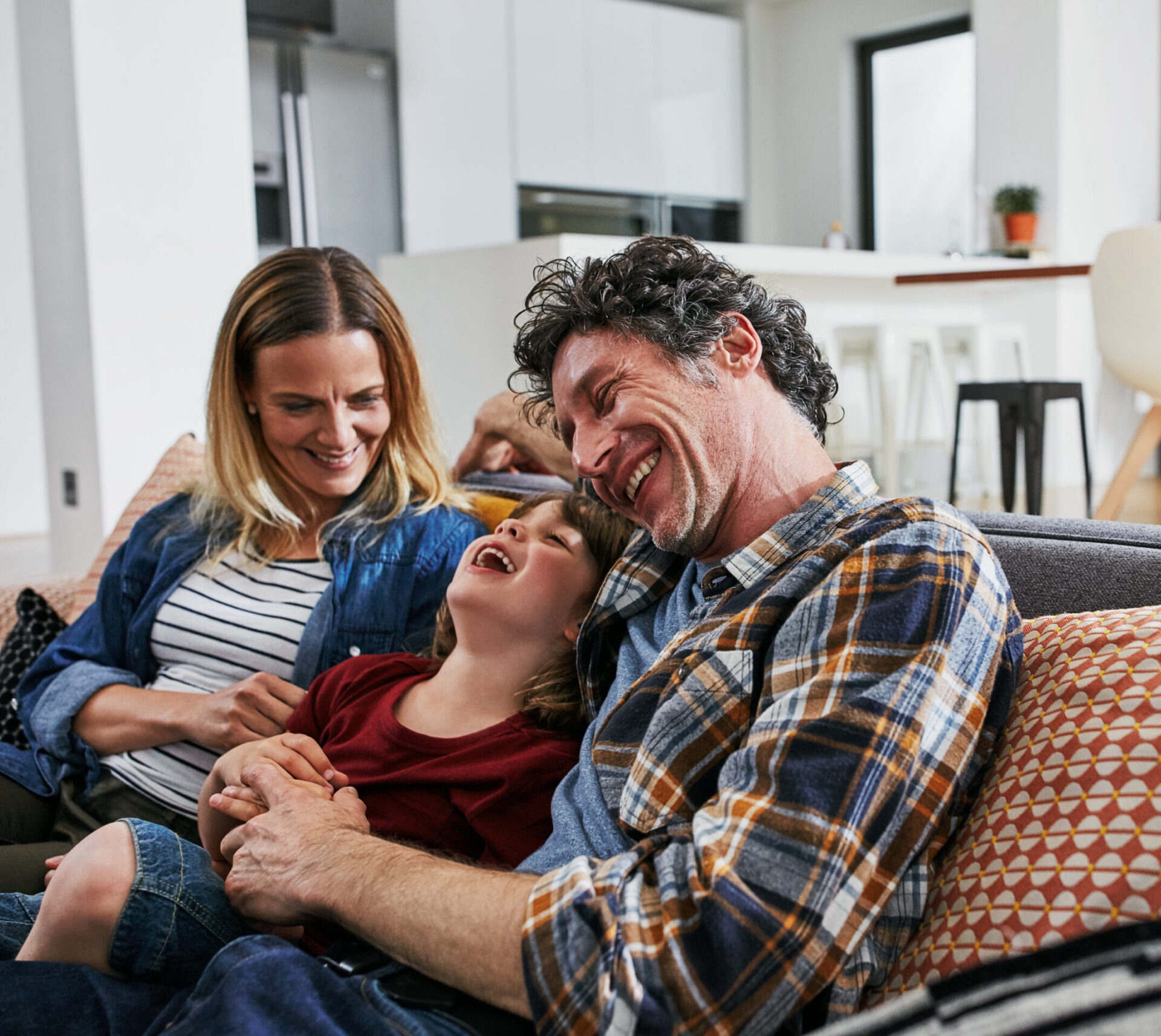 Two adults and a child are laughing and lounging on a couch in a cozy, well-lit living room, suggesting a warm, joyful family moment.