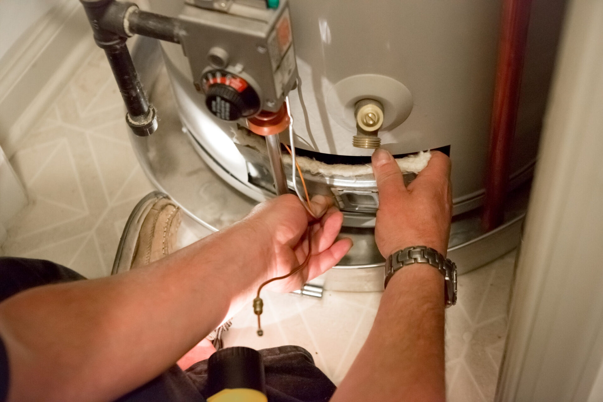 A person is servicing a water heater, adjusting components with tools. Focus on hands, wearing a watch, casual shoes visible by the unit.