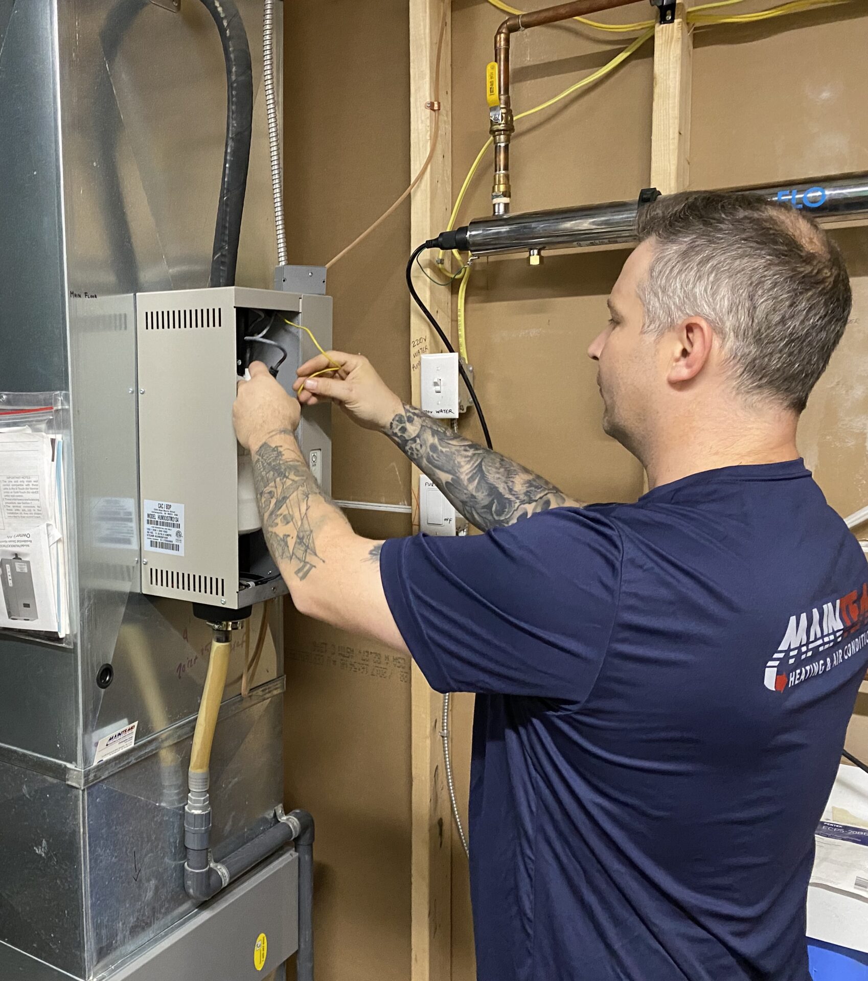 A person with tattoos is working on an electrical panel next to an HVAC system in a utility room, wearing a blue company shirt.