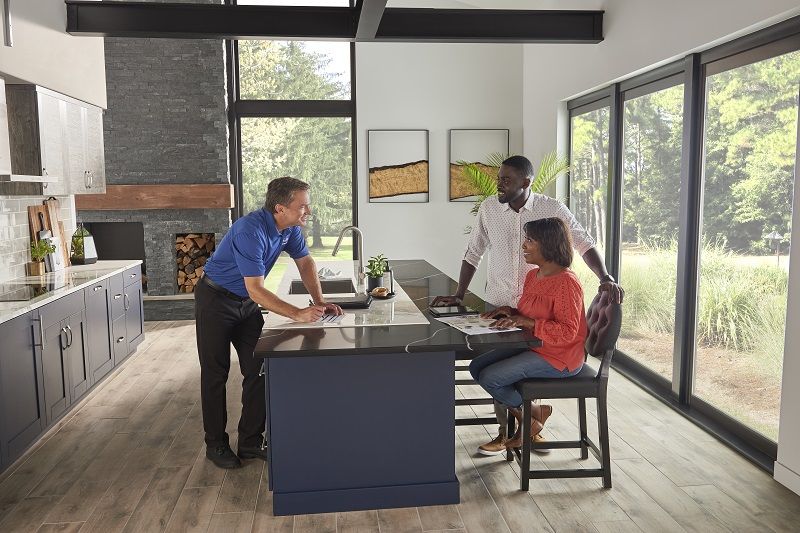 Three people are conversing in a modern kitchen with large windows, wood accents, and a center island. The environment appears relaxed and friendly.