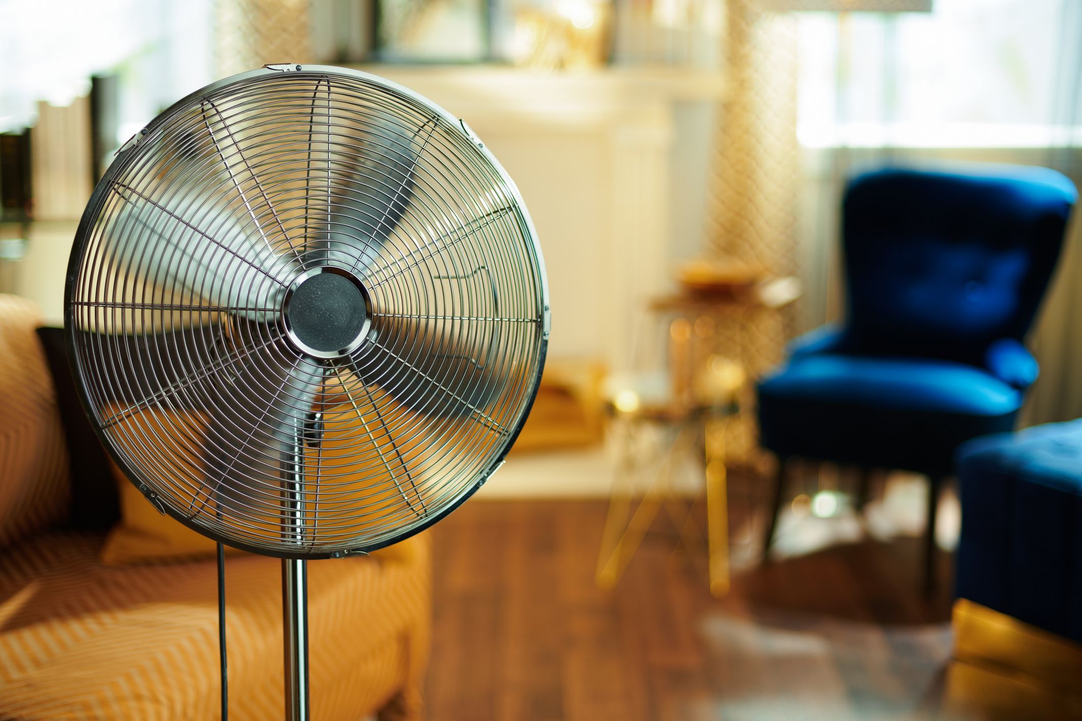 A large metal floor fan is in focus in a stylish room with blurred modern furniture, including a blue velvet chair and golden accents.