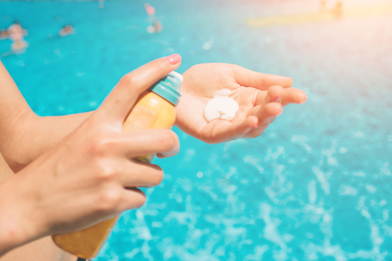 A person is applying sunscreen on their hand against a vibrant blue swimming pool backdrop. There's bright sunlight and other swimmers are visible.