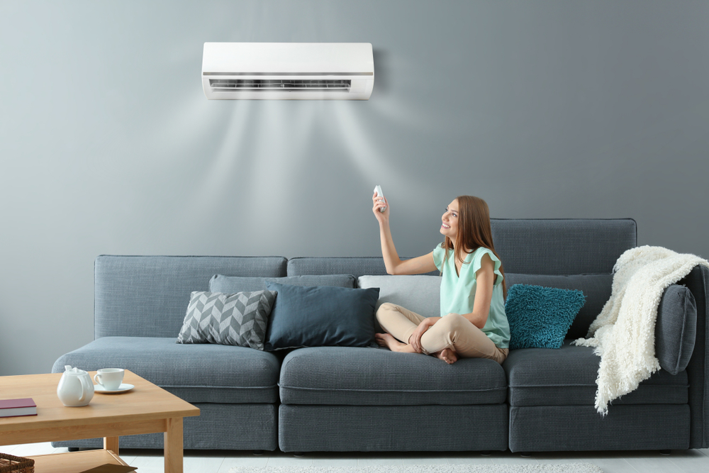 A person is sitting comfortably on a couch, using a remote to adjust an air conditioning unit on a wall in a modern, cozy living room.