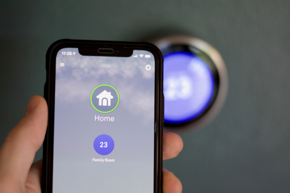 A person's hand is holding a smartphone with a smart home app opened, focused against a blurred smart thermostat in the background.