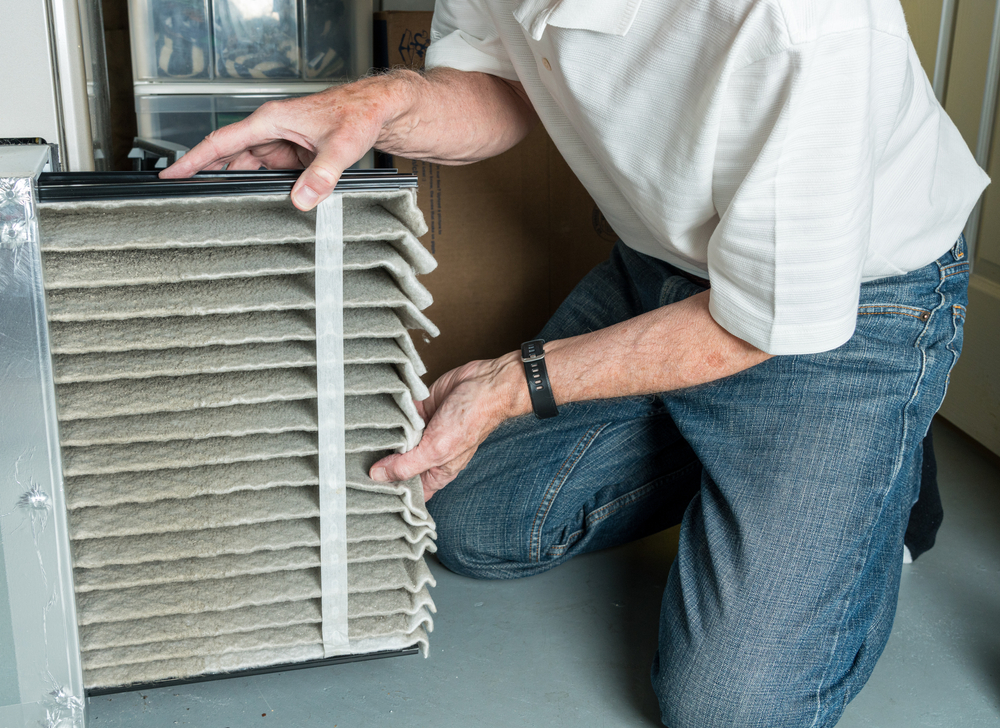 A person is kneeling and installing a folded air filter into a home HVAC unit, wearing a white shirt, jeans, and a watch.