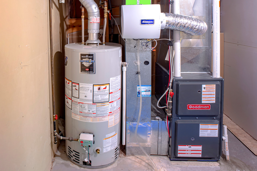 A home utility room with a gray hot water heater next to a black Goodman furnace. Pipes and ductwork are connected to both appliances.
