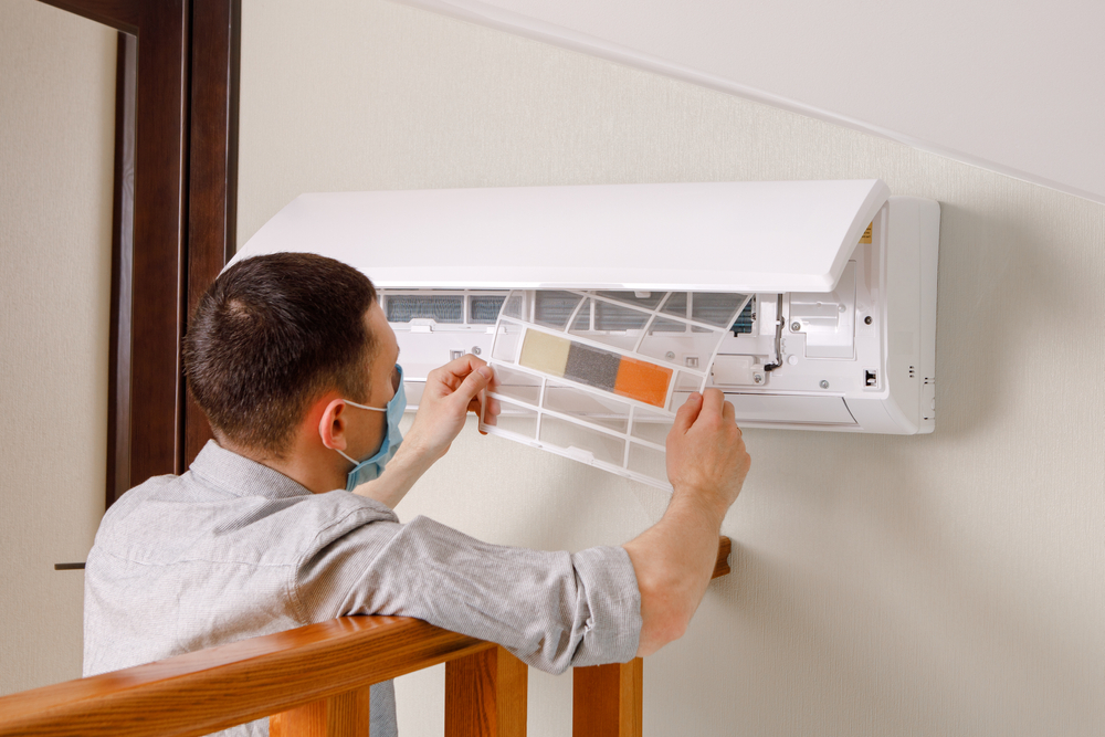 A person is maintaining an air conditioning unit on a wall, inspecting or replacing a filter, wearing casual clothes and safety glasses.