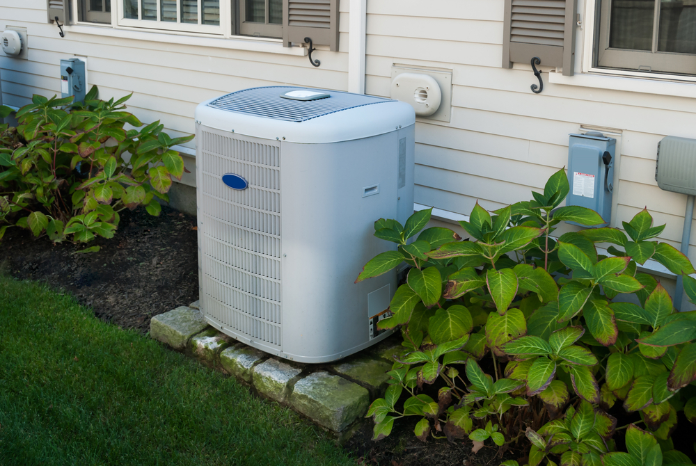 An outdoor air conditioning unit is positioned next to a house, surrounded by lush greenery, with electrical boxes on the siding nearby.