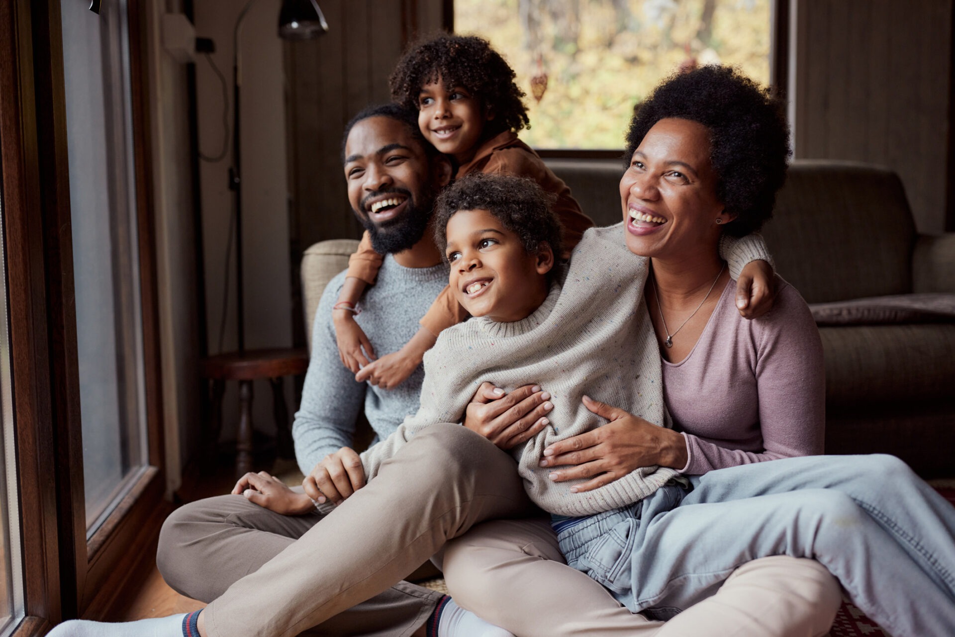 This image depicts a happy family indoors with two children. There's a person with a beard, a smiling person, and joyful kids embracing each other warmly.